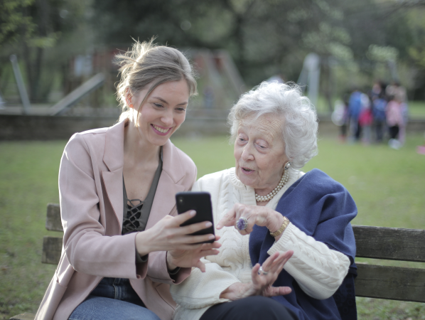 A young and an older woman are sitting on a bench in the garden and looking at a smartphone